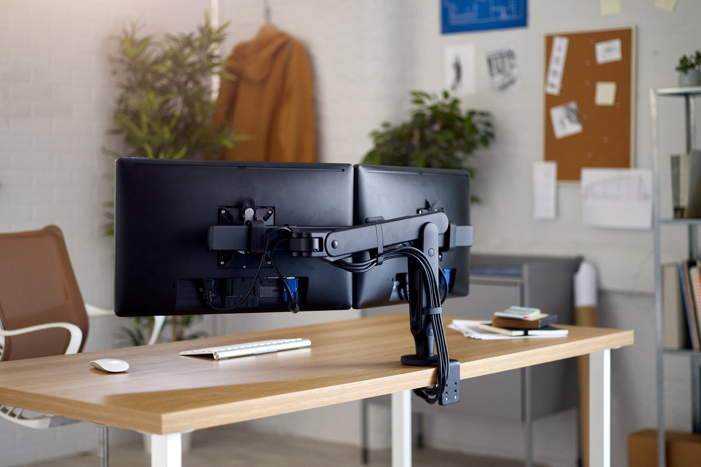 Two screens on a dual monitor arm and shared post, seen from behind, clamped to the rear edge of a timber desk with cables running down the post