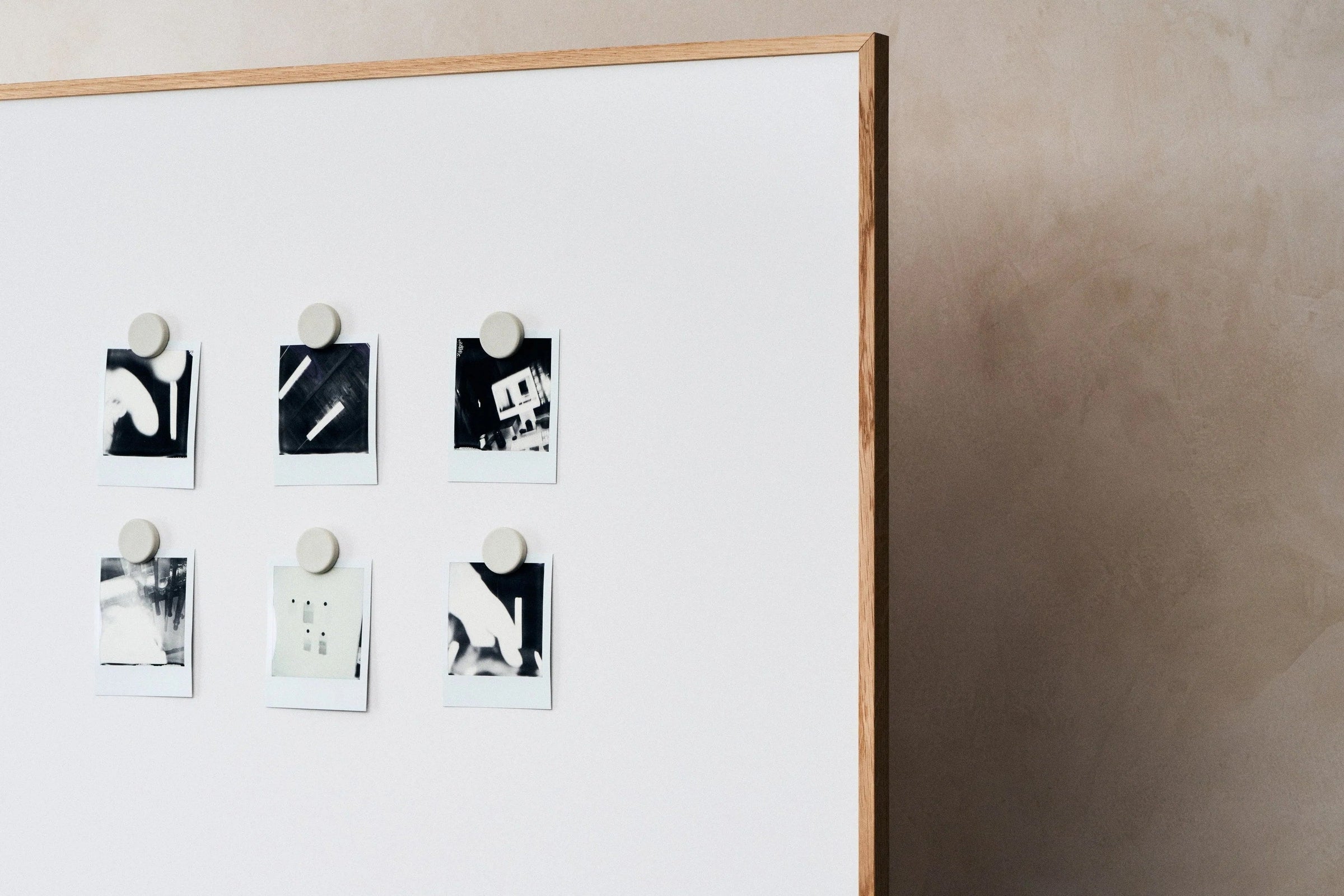 An oak framed mobile writing board standing in a workspace, its white enamelled steel surface holding six small black and white photographs under round magnets, against a plaster wall.